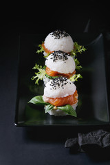 Mini rice sushi burgers with smoked salmon, green salad and sauces, black sesame served on black square plate with textile napkin over black background. Modern healthy food