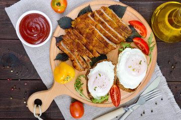 Pieces of chop (schnitzel), toast with eggs, fresh tomato on a wooden board on a dark background. Top view.