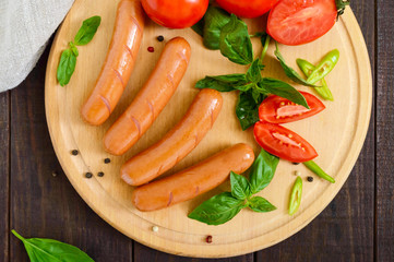 Juicy sausages with tomatoes and basil on a round cutting board on a dark wooden background. Top view.