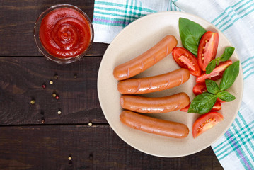 Juicy sausages with tomatoes and basil leaves on a plate on a dark wooden background. Top view.