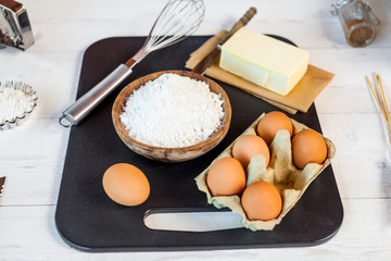 Baking ingredients in rural kitchen - dough recipe ingredients (eggs, flour, milk, butter, sugar) and rolling pin on wooden white table. Close up