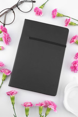 Black notebook, glasses and teapot shape plate decorated with pink carnation flowers on white background 