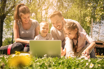 Fototapeta premium Happy family using laptop, sitting on the green grass. Family enjoying on the meadow in the spring day.