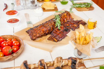 Spicy hot grilled spare ribs from a summer BBQ served with chips, corn and fresh tomatoes on an old wooden cutting board
