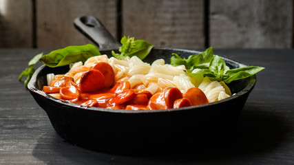 Pasta with sausages, tomato sauce and basil in a cast-iron frying pan on a tenor background