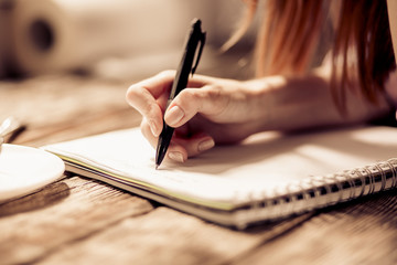 Close up view of womans hand writing with pen on notebook on wooden table. Toned image.