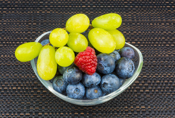 Grape bunch, blueberries, raspberry in glass bowl. Sweet juicy berries in detail. Colored fruits in oval dish on black striped placemat. Healthy fresh vegetarian snack with vitamins and antioxidants.