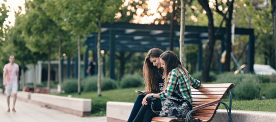Teen girls back to school bargain.Showing the elementary school supplies list.Both are eagerly waiting to see the school supply results. Happily sitting on a wooden bench, legs crossed.Back to school
