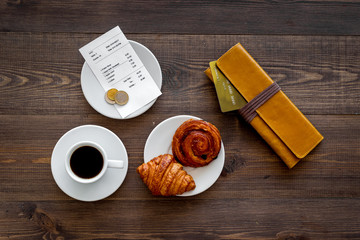 Pay bill at cafe by card. Purse, bill and bank card near coffee and croissant on dark wooden table...