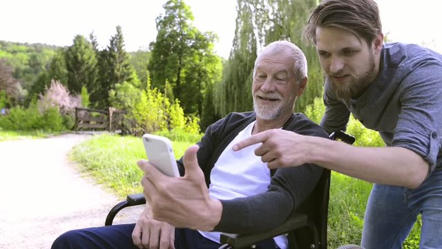 Hipster son and father in wheelchair at park taking selfie.
