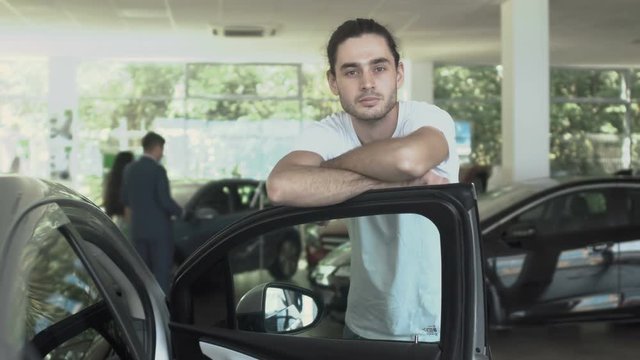 Portrait of stylish guy leans on door of modern auto in car dealership