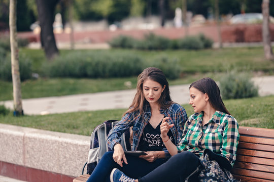 Modern girls sitting on a wooden bench. Talking seriously about their school supplies. Worried about the school supplies list. Looking at her sneakers. School backpacks are on bench. Back to school