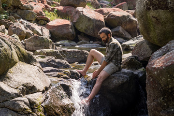 Young barefoot adult man seated on a rock on the top of little waterfall refresh himself on mountain river in the morning in summer outdoor.