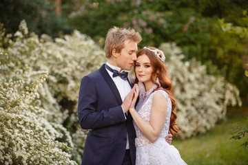 Fototapeta premium Young Bride and Groom couple in a blooming garden. Tender holding each other. Spring wedding. Redhead girl with long hairs. Young family outdoor image near blooming bush of spirea. Love and tenderness