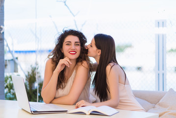 Two young pretty business-women sitting by table outdoor and working with laptop and notebook and whispering