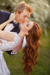 Fototapeta na wymiar Young Bride and Groom couple in a blooming garden. Tender holding each other. Spring wedding. Redhead girl with long hairs. Young family outdoor image near blooming bush of spirea. Love and tenderness