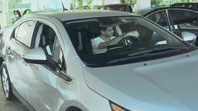 Guy waits in car cabin before car on background is going away from car showroom