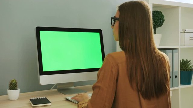 Back view portrait of young woman in the glasses and stylish shirt working on the computer with green screen in the office. Chroma key.