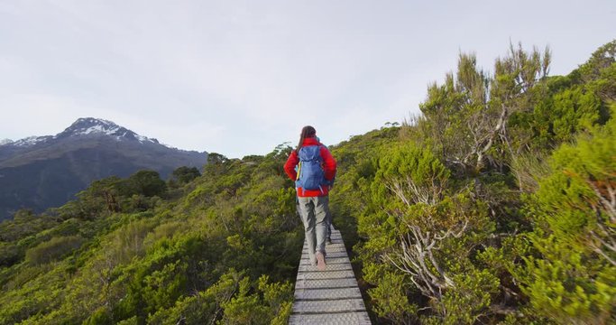 Backpackers walking on boardwalk amidst trees at Routeburn Track. Multiethnic hiking couple is tramping on footbridge at Key Summit Track. They are on vacation at Fiordland National Park, New Zealand.