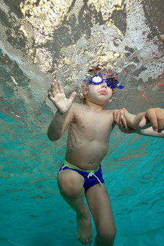 A child with glasses for swimming is learning to swim underwater in the bubbles. Bottom view from under the water. Portrait. Vertical orientation