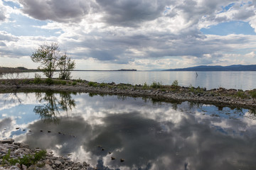 A dock in a lake, with a beautiful, deep blue sky with white clouds reflecting on water, along with some trees