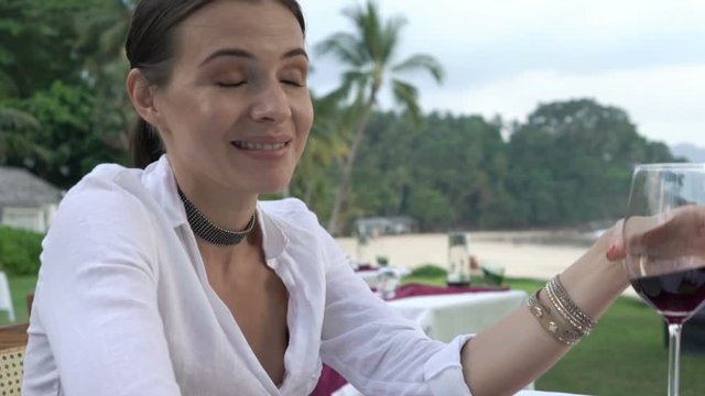 Young, happy woman talking to camera and drinking wine in beach restaurant
