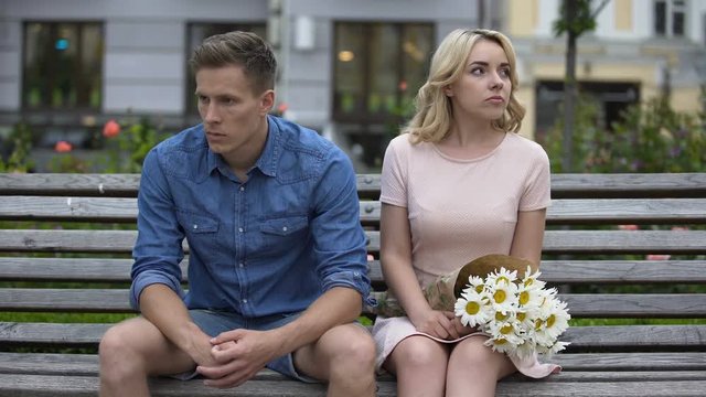 Unhappy couple sitting after fight, girl leaving and throwing flowers on bench