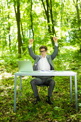 Fototapeta premium Portrait of young handsome succesful business man in suit working at laptop at office table with raised hands of victory in green forest park. Business concept.