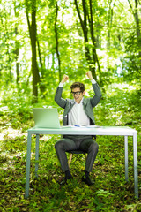 Portrait of young handsome succesful business man in suit working at laptop at office table with raised hands in green forest park. Business concept.