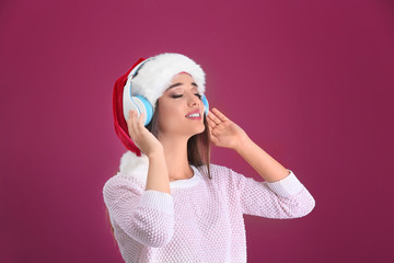 Beautiful young woman in Santa hat listening to Christmas music on color background