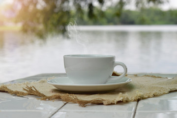 hot coffee in white cup on table with lake and trees nature background.
