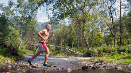 A young sporty bald man with a naked torso and short white shorts running down a mountain river on...