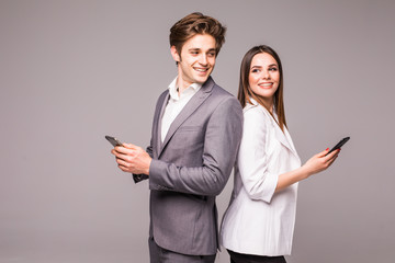 Young couple is using smart phones and smiling while standing back to back on a gray background. Look each other.