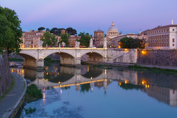 Saint Angel bridge and Saint Peter Cathedral with a mirror reflection in the Tiber River during morning blue hour in Rome, Italy.