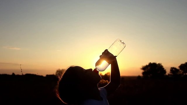 Silhouette of young girl drinking water from bottle in wheat field at sunset in summertime. Rehydrate your body.