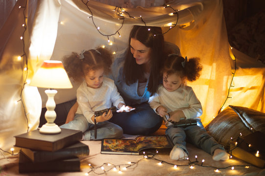 Reading and family games in children's tent. Mother and two twins daughters with books and flashlight before going to bed.