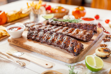 Spicy hot grilled spare ribs from a summer BBQ served with chips, corn and fresh tomatoes on an old wooden cutting board