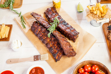 Spicy hot grilled spare ribs from a summer BBQ served with chips, corn and fresh tomatoes on an old wooden cutting board