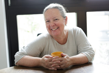 Fröhliche Seniorin sitzt mit Tasse am Tisch zuhause
