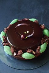 Contemporary Chocolate, Cherry and Pistachio Layered Mousse Cake glazed with chocolate mirror glaze, on dark background.