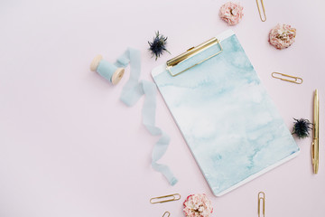 Flat lay of clipboard, rose buds, blue ribbon, golden pen and clips on pale pink background. Top view stylish decorated mock up.