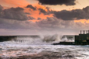 Storm waves crash over break water under sunset sky