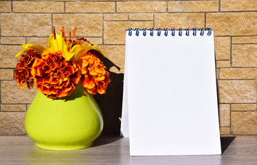 Mock up, abstract background, blank white sheet of paper in the notebook and yellow ceramic vase with a bouquet of orange flowers against a brick wall