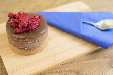 Piece of cake with chocolate glaze and raspberries on a plate on a wooden background. Blue napkin with a spoon beside the cake.
