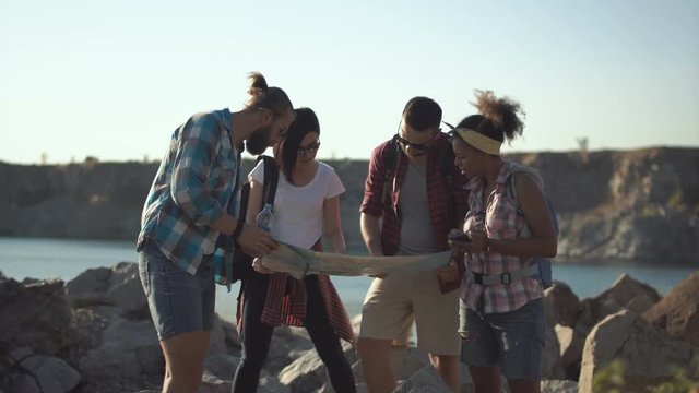 Young people friends with backpacks on rocky coastline on lake trying to find right way using map and smartphone with gps.