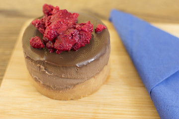 Piece of cake with chocolate glaze and raspberries on a plate on a wooden background. Blue napkin with a spoon beside the cake.