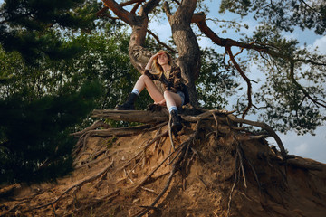 Attractive tourist girl sitting on root of tree and taking rest after walk
