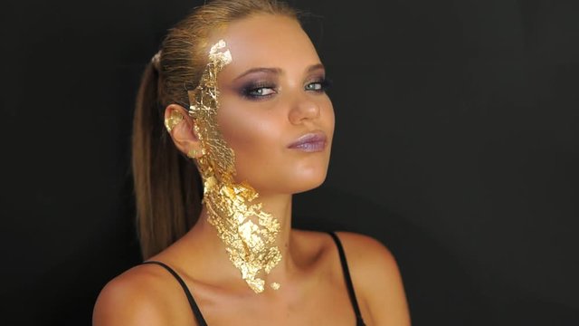 Fashion model standing against the black wall and striking a pose for studio fashion photo shoot. stylish golden makeup and hairstyle. Skin on face and neck covered with golden metallic pieces