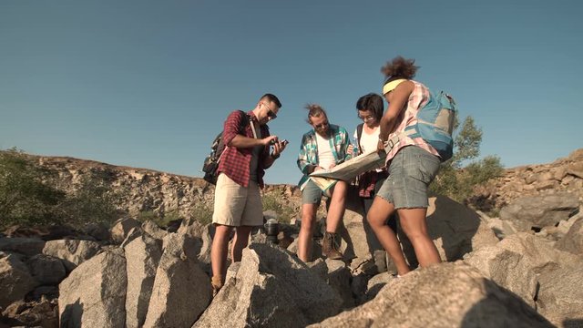 Young diverse mixed ethnic people travelers with backpacks on rocky coastline exploring map using gps tracker.