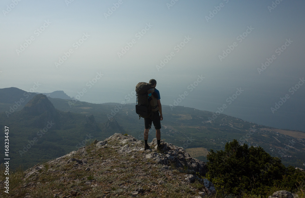 Wall mural A young tourist with a backpack stands at the edge of the cliff and looks at the mountain landscape and the sea.